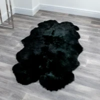 A large black rug with an organic shape lies on a light grey wooden floor in a minimalist room. A Natural White Shorn Sheepskin Rug adds contrast near white walls and a metal-legged table partially visible in the corner.