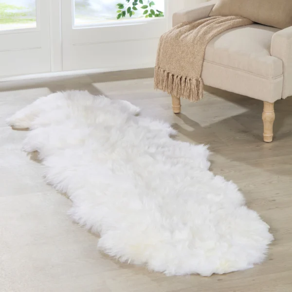 A Natural White Double Sheepskin Rug rests on a light wooden floor beside a beige armchair with a tan fringed throw, as large windows fill the space with light and showcase the greenery outside.