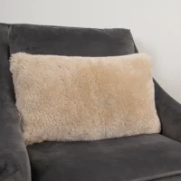 A rectangular, fluffy cream-coloured sheepskin cushion with a soft sheepskin texture rests on a charcoal grey, suede-like armchair against a plain white wall. The sheepskin cushion contrasts beautifully with the dark chair, enhancing the cosy and inviting atmosphere.