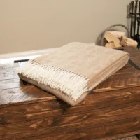 A beige wool throw blanket with a subtle diamond pattern and white fringe is neatly folded on a rustic wooden chest. Firewood and fireplace tools in the background create an inviting, homely setting with warmth and charm.