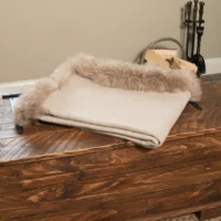 The Sand Beige Wool Throw with Sheepskin Trim is folded atop a rustic wooden chest, whilst firewood, fireplace tools, and a brick hearth in the background complete the cosy scene.