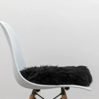 A side view of a modern white plastic chair with wooden legs and metal supports, topped with a Midnight Black Round Sheepskin Cushioned Seat Pad. The background is a plain light grey wall, giving the scene a minimalist look.