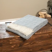 A folded light blue wool blanket with a subtle geometric pattern and white fringe rests on a rustic wooden chest. Firewood, metal fireplace tools, and a light beige wall are visible in the cosy background.
