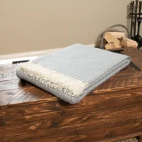A folded, light blue and white fringed wool blanket rests on a rustic wooden chest with visible wood grain and metal accents. In the background, a stack of firewood and a cosy fireplace tool set sit against a beige wall.