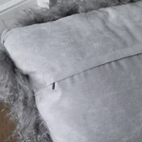 Close-up of a soft, light grey suede pillow with a flap-style opening. The pillow is bordered by fluffy, textured sheepskin material, adding a cosy and luxurious appearance. In the background, there’s a hint of wooden floor and white wall trim.