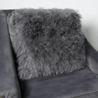 A square, fluffy, dark grey pillow made from real sheepskin with a shaggy, curly texture rests on a matching dark grey suede armchair. The pillow’s soft, long fibres give it a cosy and inviting appearance against the smooth microfiber fabric of the chair.