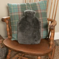 A grey sheepskin hot water bottle rests against a green and blue plaid cushion on a wooden rocking chair with curved armrests, creating a cosy, inviting corner by a beige wall.