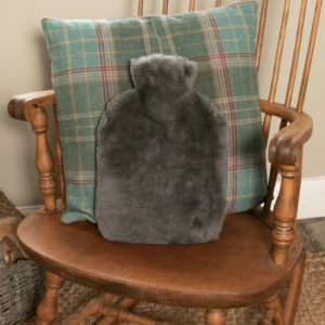 A grey sheepskin hot water bottle rests against a green and blue plaid cushion on a wooden rocking chair with curved armrests, creating a cosy, inviting corner by a beige wall.