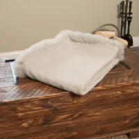 A folded, Grey Linen Throw with Sheepskin Trim. Stacked firewood and fireplace tools are seen in the background against a neutral wall.