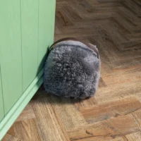 The Battleship Grey Sheepskin Door Stop, featuring a brown handle and soft, dense sheepskin, rests against a light green door on a herringbone wooden floor, its plush texture contrasting with the door’s smooth surface.