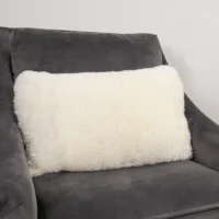 A fluffy, rectangular white sheepskin wool cushion with a soft texture rests on a dark grey, velvety armchair. The plain, light-coloured wall in the background highlights the cushion’s plush appearance through contrasting tones.