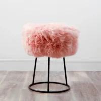 A sheepskin stool with pink, fluffy upholstery sits on a round black metal frame with four slim legs, placed on a light wood floor against a plain white wall.