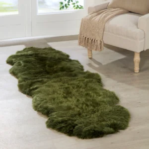 A Green Double Sheepskin Rug with an organic shape lies on a light wood floor beside a beige upholstered chair draped with a tan fringed throw, as sunlight streams in through large windows.