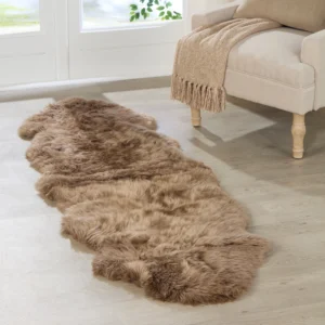 A Brown Double Sheepskin Rug rests on a light wooden floor in front of a sofa with a beige fringed throw, as large windows in the background let in natural light.