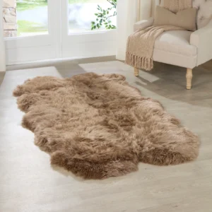 A Brown Quad Sheepskin Rug lies on a light wood floor in a bright room. Behind it, a beige armchair with a cushion and folded throw is placed near tall glass doors that overlook greenery outside.