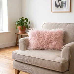 A rectangular, fluffy pink sheepskin cushion with a curly fur texture rests on a beige linen-look armchair. The chair features plush cushioning and armrests, set against a plain white wall background.
