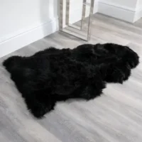 A Midnight Black Sheepskin Rug – Extra Large with irregular edges lies on a light grey wood floor near a white wall and a modern silver table leg. Its fluffy texture contrasts strikingly with the smooth, hard surface beneath.