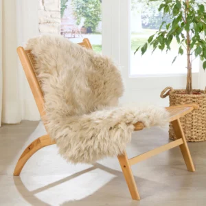 A wooden chair with a woven cane seat is draped with an Extra Large Champagne Sheepskin Rug. The chair sits by a sunny window on a light wood floor, with a potted green plant in a basket nearby, creating a cosy atmosphere. Keywords: sheepskin.