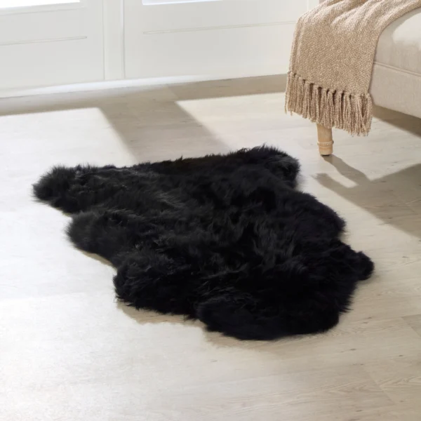 An extra large black sheepskin rug lies on a light wooden floor beside a beige sofa with wooden legs. A tan knitted throw blanket is draped over the sofa arm as sunlight streams through large windows, brightening the room.