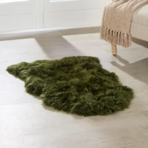A soft, irregularly-shaped dark green rug lies on a light wood floor. In the upper right, a beige sofa with wooden legs and a tan fringed throw sits nearby. An Extra Large Green Sheepskin Rug adds cosy style. White window panels are in the background.