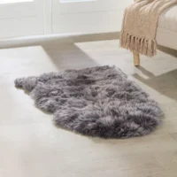 A plush, grey Grey Sheepskin Rug – Extra Large rests on a light wooden floor by a beige sofa with wooden legs, draped with a tan fringed knitted blanket. Sunlight pours in through large windows in the background.