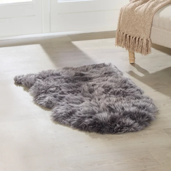 A plush, grey Grey Sheepskin Rug – Extra Large rests on a light wooden floor by a beige sofa with wooden legs, draped with a tan fringed knitted blanket. Sunlight pours in through large windows in the background.