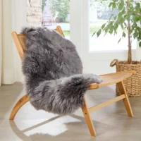 A wooden lounge chair with a woven back and seat is draped with a thick, fluffy Grey Sheepskin Rug – Extra Large. The chair sits on a light wood floor by a large window, with sunlight streaming in and a leafy potted plant in a woven basket nearby.