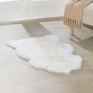 An extra large White Sheepskin Rug lies on a light wooden floor beside a beige sofa with wooden legs, draped with a tan knitted throw blanket. Sunlight streams through tall windows in the background.