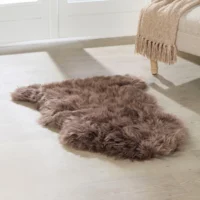 An extra large, soft taupe sheepskin rug with a wavy, fluffy texture rests on a light wooden floor next to a beige sofa draped with a fringed tan throw. Sunlight pours in through white glass doors.