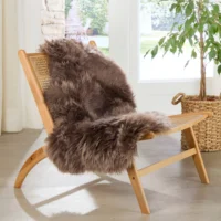 A wooden chair with a woven cane seat and back is draped with an Extra Large Taupe Sheepskin Rug. The chair stands on a light-coloured floor near a window and a leafy potted plant in a woven basket, bathed in sunlight.