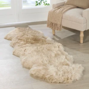 A Champagne 2 Pelt Sheepskin Rug lies on a light wood floor by a large window. Next to it, a cream armchair with wooden legs displays a tan fringed throw. Green leaves can be seen outside the window.