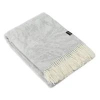 The Light Grey Wool Blanket - Dragon Tree Leaf features soft, folded wool with a cream-coloured fringe on one edge and a small black label near the fringe. Plush and neatly arranged, it is displayed against a white background.