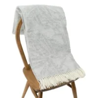 The Light Grey Wool Blanket - Dragon Tree Leaf, featuring a white fringe, hangs over a simple wooden chair with its plush ends draped on the seat, set against a plain white background.