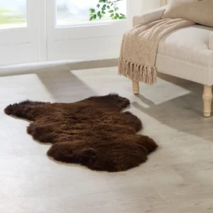 A Brown British Sheepskin Rug rests on light wood flooring by a beige sofa with wooden legs, draped with a textured beige fringed throw, as sunlight from large windows highlights greenery outside.