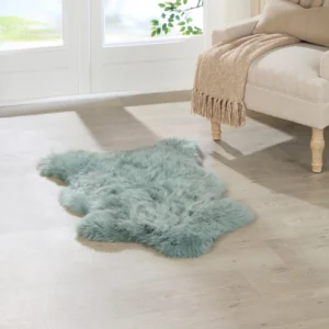 A Mint Green British Sheepskin Rug lies on a light wood floor near a cream sofa with wooden legs. A beige, fringed throw blanket is draped over the sofa arm, whilst sunlight streams through large glass doors in the background.