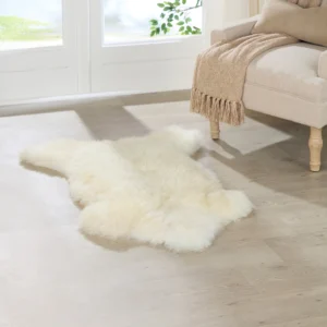 An Ivory White British Sheepskin Rug rests on a light wood floor beside a beige armchair with a tan knitted throw. Sunlight from large windows with sheer curtains casts soft shadows, highlighting the sheepskin rug in this cosy, serene room.