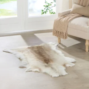 A Light Coloured Reindeer Hide rug rests on a light wood floor near a beige sofa with a tan fringed throw. Sunlight pours through large glass doors, revealing green foliage outside.