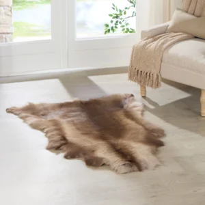 A Dark Coloured Reindeer Hide rug is placed on a light wooden floor in front of a sofa with a tan, fringed throw. Sunlight from large glass doors illuminates greenery outside, making the space feel cosy and inviting.