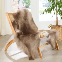 A wooden lounge chair with a woven backrest is draped with a dark-coloured reindeer hide. Beside it, a potted green plant in a woven basket rests on the light-coloured floor near a window with white curtains.