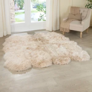 A Champagne Octo Sheepskin Rug lies on a light wood floor in a bright room. An upholstered armchair draped with a beige throw sits by tall windows with sheer white curtains, framing views of outdoor greenery.