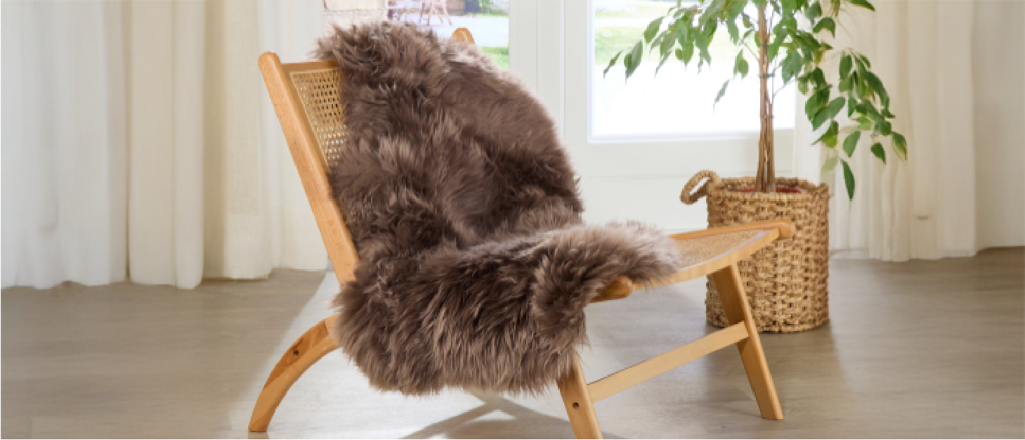 A wooden lounge chair with a woven cane back is draped with a fluffy grey sheepskin rug. Behind the chair, a woven basket holds a leafy green plant. Light filters through sheer curtains and a glass door, creating a cosy, inviting atmosphere.
