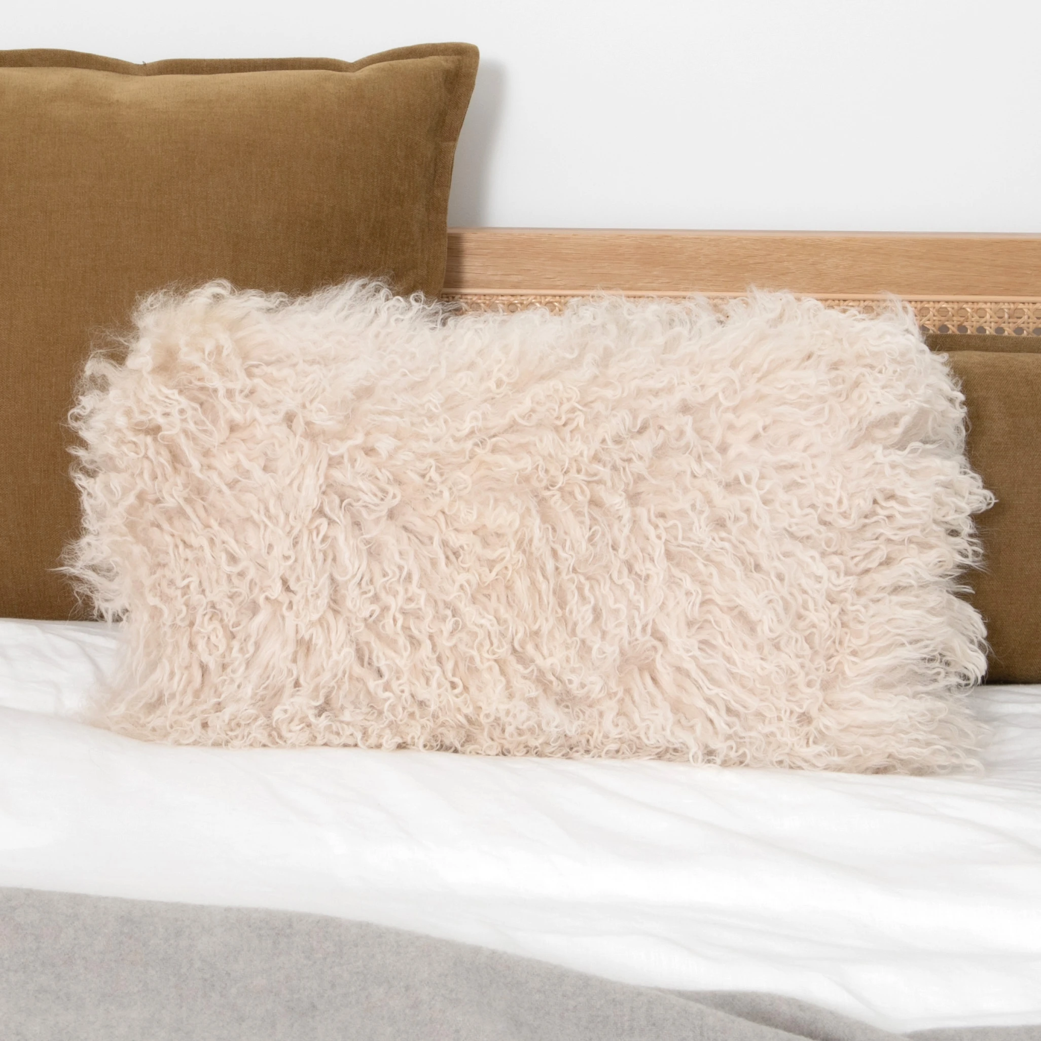 Rectangular cream Mongolian sheepskin cushion with long curly pile styled on a bed in front of a brown velvet pillow.