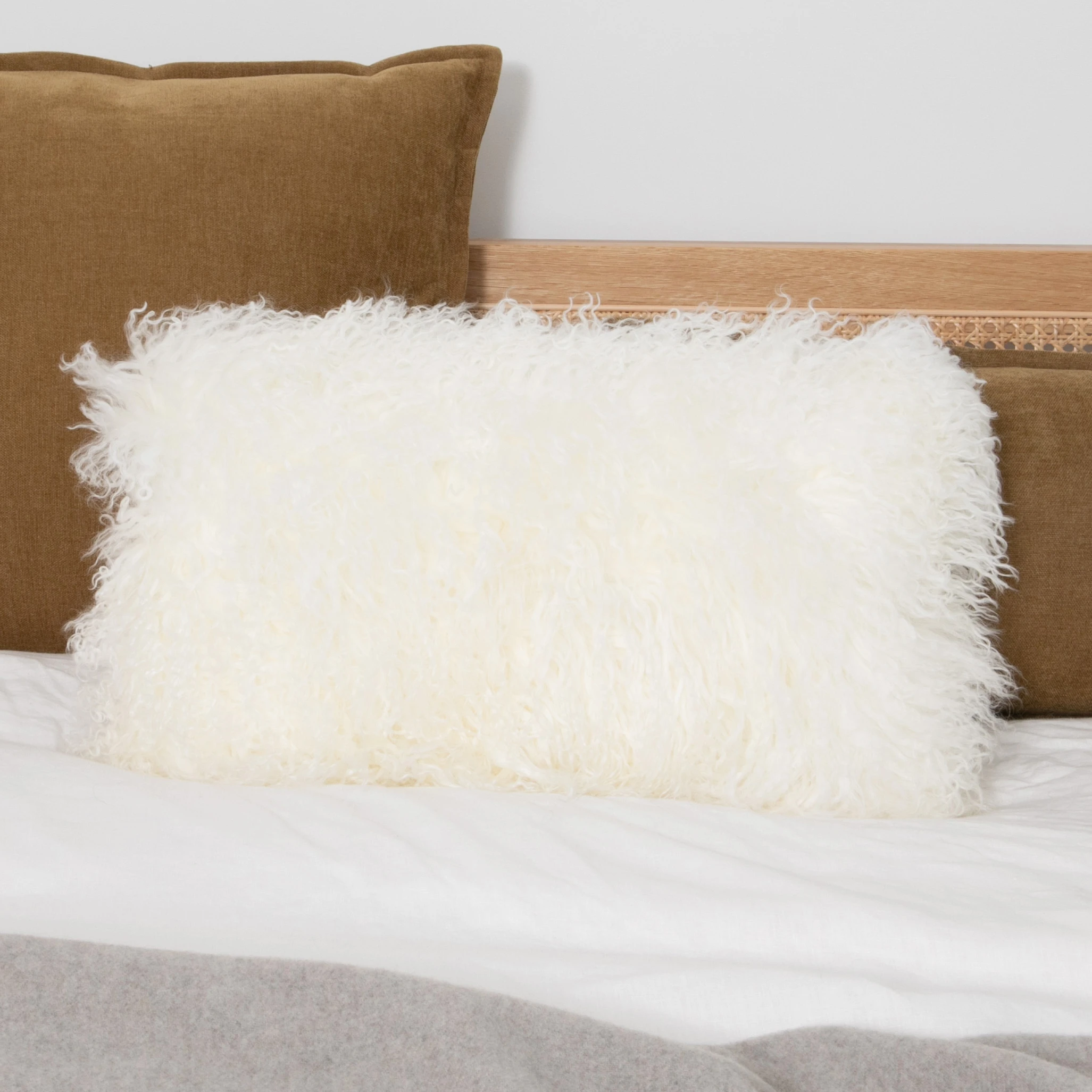 White rectangular curly sheepskin cushion styled on a bed with brown velvet pillows.