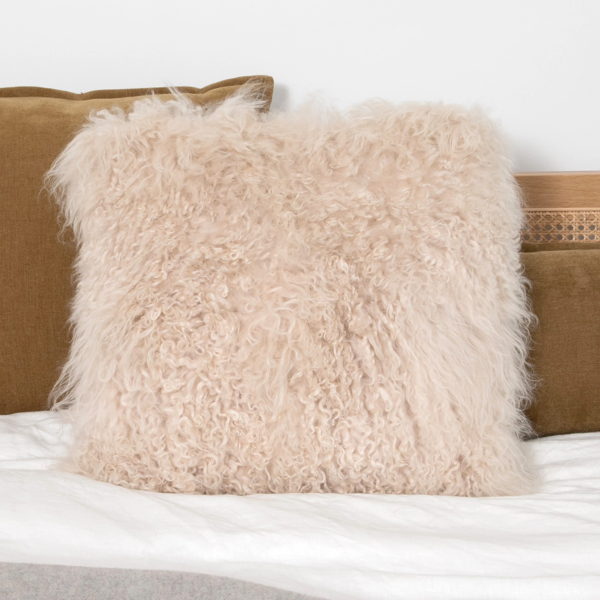 Square blush Mongolian sheepskin cushion with long curly pile on a white bed against a tan cushion and wooden headboard.