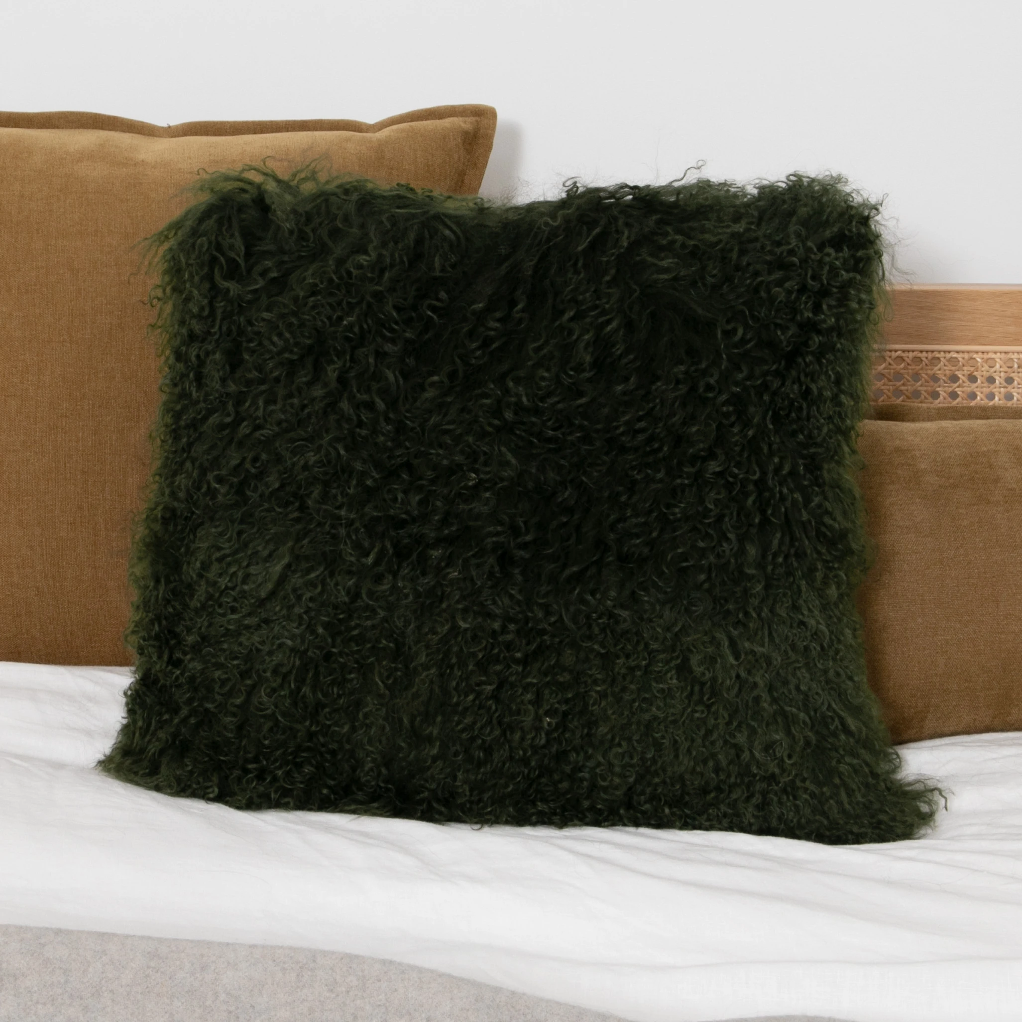 Dark forest green curly Mongolian sheepskin cushion styled on a bed with tan velvet pillows behind it.