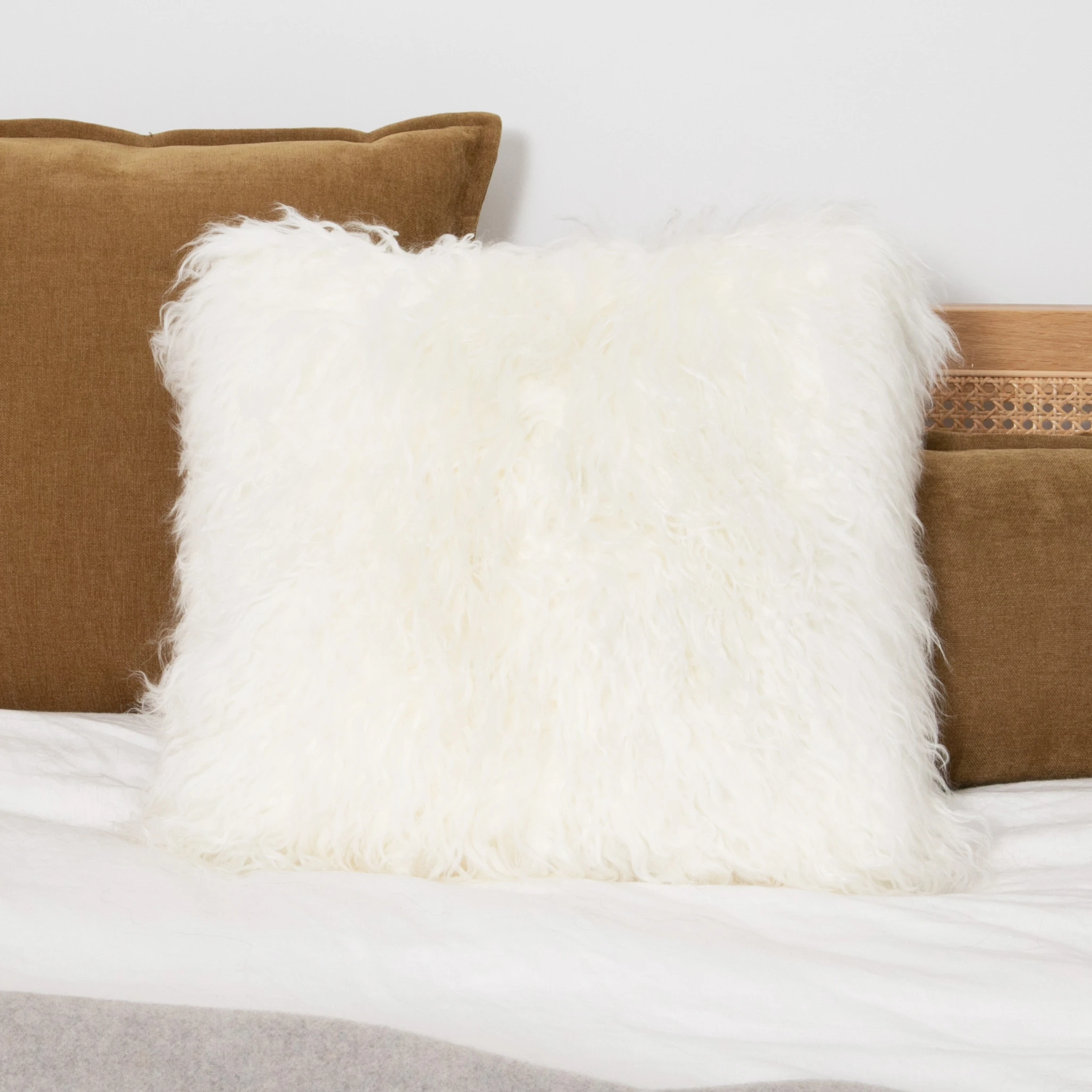 Square white Mongolian sheepskin cushion with long curly pile styled on a bed with brown cushions behind.